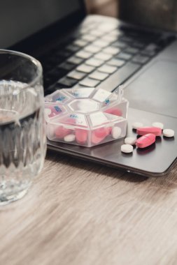 Organizer weekly shots with glass of water on workplace background and laptop Closeup of medical pill box with doses of tablets for daily take medicine with white pink drugs and capsules. Medication