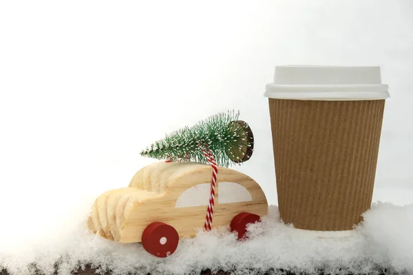 Wooden car carrying Christmas tree with paper cup mock up of coffee or hot chocolate over snow. Copy space for text Toy car in snowy landscape. Merry Christmas and Happy New Year concept. Winter is