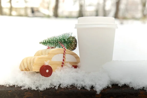 Wooden car carrying Christmas tree with paper cup mock up of coffee or hot chocolate over snow. Copy space for text Toy car in snowy landscape. Merry Christmas and Happy New Year concept. Winter is