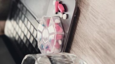 Düzenleyici haftalık çekimlerini iş arka planında bir bardak suyla dikey olarak büyüt ve laptopta tıbbi hap kutusunu günlük dozda haplarla kapat beyaz pembe ilaçlarla ilaç al ve