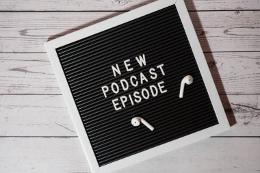 Yeni PODCAST EPISODE yazılı duyuru panosu ahşap masada kablosuz kulaklıklarla. Podcast konsepti. Radyodaki ses iyileştirme sağlık alışkanlıkları.