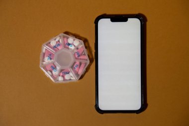 Mobile phone with white screen copy space template medical pill box with doses of tablets for daily take medicine with white pink drugs and capsules. Daily vitamins at home. Medication dietary