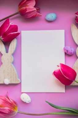 Beyaz boş zarf paskalya yumurtalarını ve menekşe rengi pastel arka planda pembe laleli ahşap tavşanları kopyalıyor. Tebrik kartı. Düz kompozisyon üst görünümü. Dekorasyon minimalist modern tasarım