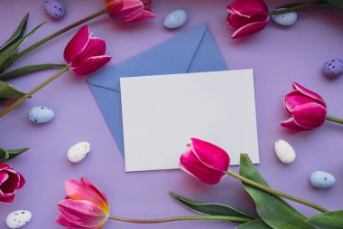 Beyaz boş zarf, menekşe renginde pembe laleleri olan Paskalya yumurtalarını kopyalıyor. Tebrik kartı. Düz kompozisyon üst görünümü. Dekorasyon minimalist modern tasarım şablonu. Çiçek.