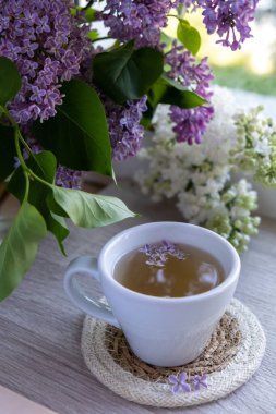 Lezzetli siyah çay beyaz bardakta pencere kenarında aromatik leylak çiçekleriyle. Bahar kompozisyonu bir bardak leylak çayı içerken mor leylak dalları çiçek açıyor. Kopya alanı tebrik kartı için natürmort hayat