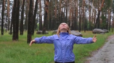 Ağır çekimde mavi yağmurluklu mutlu genç kadın parktaki ormanın tadını çıkarıyor. Yağmurlu hava tahminlerinde gerçek duygular açık havada kollarını açar. Turist istirahati ve özgürlük. Sonbahar mevsimi