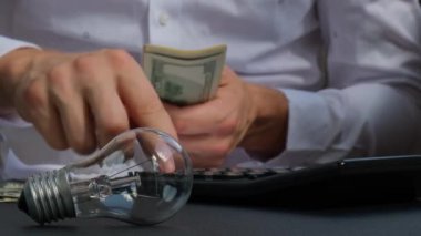 Elektrik ampulü. Adam, elektrik banknotlarındaki harcamaları hesaplayan hesap makinesindeki dolar hesaplarını sayıyor. Tanınmayan bir iş adamının eli kulağında. Enerji için yüksek fiyatlar. Bütçeyi biriktir