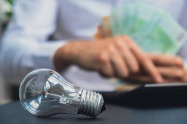 Elektrik ampulü. Adam, hesap makinesindeki euro nakit hesaplarının elektrik banknotlarının faturalarını sayıyor. Tanınmayan bir iş adamının eli kulağında. Enerji için yüksek fiyatlar. Biriktir
