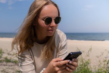 Güneş gözlüklü, deniz kabuğu bilezikli, elinde akıllı telefon tutan genç bir kadın. Sahilde selfie çekmek için cep telefonu kullanan nüfuzlu biri. Sahilde el sallıyor. Tatil reklamı konsepti. Sosyal