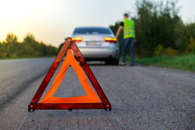 Yansıtıcı yelek giymiş, tanınmaz halde üzgün bir sürücü. Kaputu açık kırık bir arabanın yanında duran erkek sürücü. Kırmızı üçgen diğer yol kullanıcılarını araba arızası ya da motor arızası konusunda uyarmak için kırsal bölgede durur
