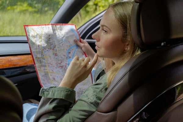 Blonde woman sitting inside the car checking the route on map to get to the destination. Afraid to get lost in forest. Young tourist explore local travel making candid real moments. True emotions