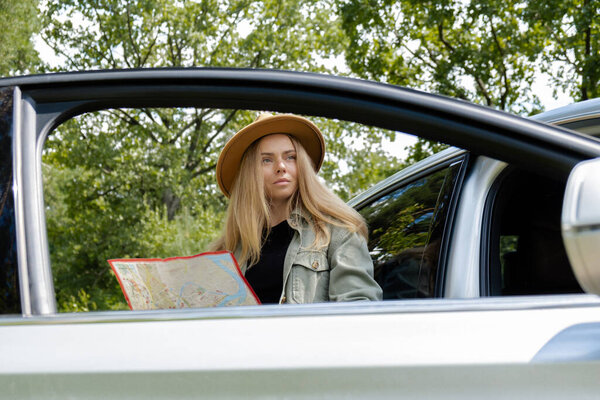Smiling young woman in hat stoped on road to check the route on travel map. Local solo travel on weekends concept. Exited woman explore freedom outdoors in forest. Unity with nature lifestyle, rest