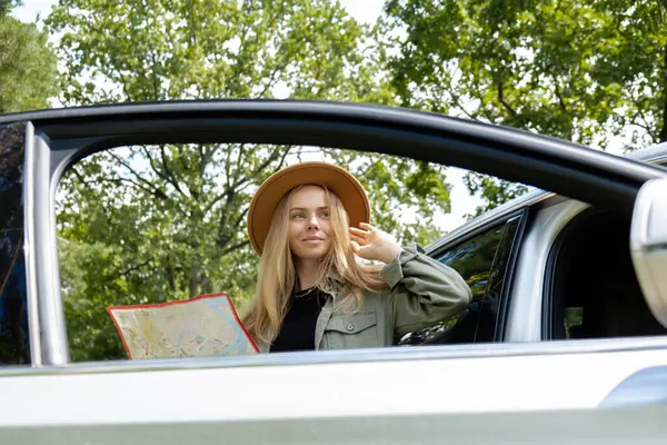 Blonde woman in hat staying next to car door checks the route on map to get to the destination. Young tourist explore local travel making candid real moments. True emotions expressions of getting away