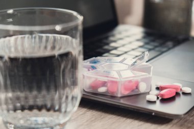 Organizer weekly shots with glass of water on workplace background and laptop Closeup of medical pill box with doses of tablets for daily take medicine with white pink drugs and capsules. Medication