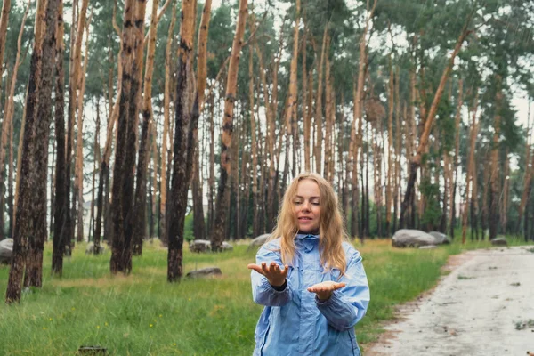Overjoyed blonde woman in blue raincoat enjoying silence natural green environment woods in the forest. Happy emotion open arms outdoors in rainy weather. Unity with nature physical mental wellbeing