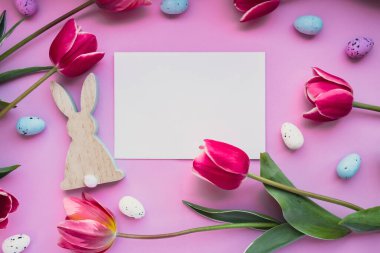 Beyaz boş zarf paskalya yumurtalarını ve menekşe rengi pastel arka planda pembe laleli ahşap tavşanları kopyalıyor. Tebrik kartı. Düz kompozisyon üst görünümü. Dekorasyon minimalist modern tasarım