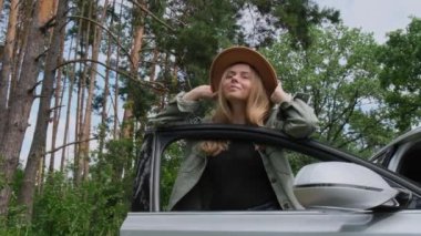 Şapkalı, gülümseyen genç bir kadın, yolda durup arabayı durdurdu. Hafta sonları yavaş çekim yerel solo seyahat kavramı. Çıkan kadın ormanda özgürlüğü keşfediyor. Doğayla birlik