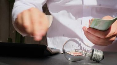Elektrik ampulü. Adam, hesap makinesindeki euro nakit hesaplarının elektrik banknotlarındaki harcamaları sayıyor. Tanınmayan bir iş adamının eli kulağında. Enerji için yüksek fiyatlar. Bütçeyi biriktir