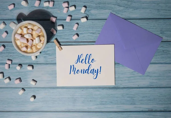 HELLO MONDAY blue monday concept text violet envelope with white cup of coffee and marshmallows on wooden background. The most depressing day of the year in January. Day commit suicide and depression motivation sign. Top view, flat lay
