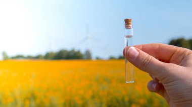 Kanola çiçeklerinin çiçek tarlasının arka planında sıvı bulunan el ele tutuşan test tüpü. Doğal arka planda kolza yağı. Tüp kimyasal analiz ph testinde çiftçi toprak örneği. Agrokimyasal analiz toprak kalitesi izleme konsepti. Rüzgar türbinleri o