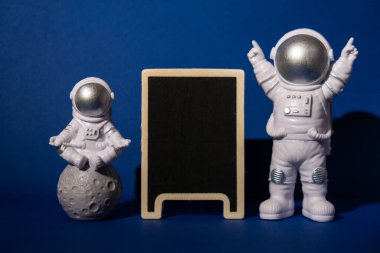 Plastic toy astronaut with blackboard template mock up for your text on colorful blue background Copy space. Concept of out of earth travel, private spaceman commercial flights. Space missions and Sustainability