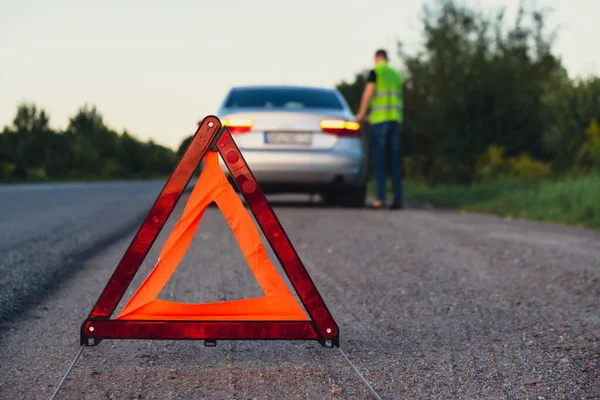Kırık gümüş rengi lüks araba acil durum kazası. Adam sürücü yola kırmızı üçgen dur işareti yerleştiriyor. Spor otomobiller yolda sinyal lambalarını çalıştırdı. Güvenlik prosedürü