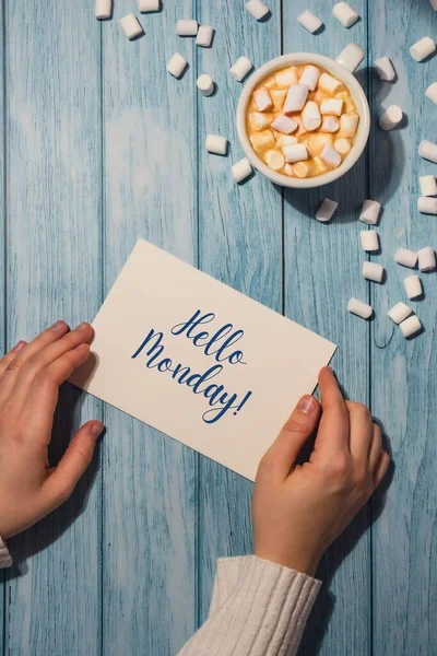 HELLO MONDAY blue monday concept text Female hands holding card with white cup of coffee and marshmallows on wooden background. The most depressing day of the year in January. Day commit suicide and depression motivation sign. Top view, flat lay