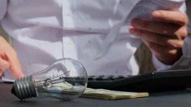 Elektrik ampulü. Adam, elektrik banknotlarının faturalarını, hesap makinesindeki dolar hesaplarını sayıyor. Tanınmayan bir iş adamının eli kulağında. Enerji için yüksek fiyatlar. Biriktir