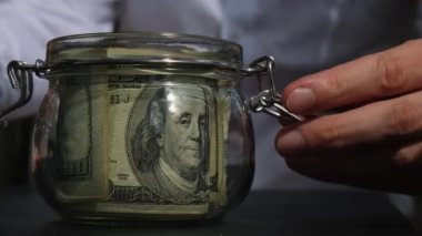 Adam kumbaradaki cam kavanozdan gelen dolar banknotlarını sayıp sayıyor. Tanınmayan bir iş adamının eli kulağında. Bütçe yatırımı kavramını sakla. Amerikan doları.