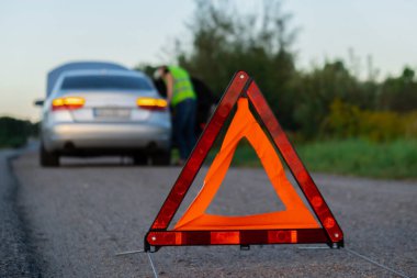 Yansıtıcı yelek giymiş, tanınmaz halde üzgün bir sürücü. Kaputu açık kırık bir arabanın yanında duran erkek sürücü. Kırmızı üçgen diğer yol kullanıcılarını araba arızası ya da motor arızası konusunda uyarmak için kırsal bölgede durur