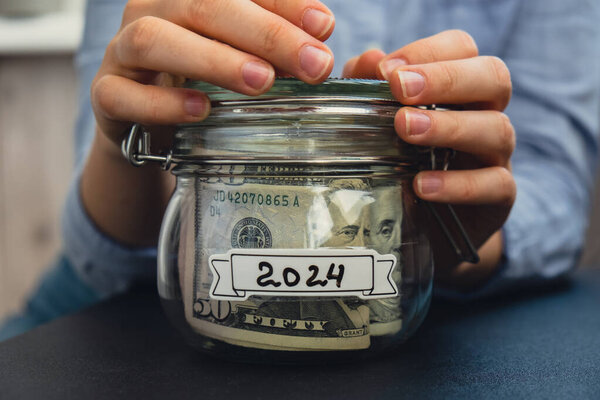 Unrecognizable woman holding Saving Money In Glass Jar filled with Dollars banknotes. 2024 transcription in front of jar. Managing personal finances extra income for future insecurity background
