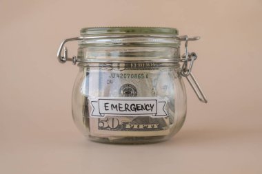 Dolarla dolu cam kavanozda para biriktiriyorum. EMERGENCY transkripti kavanozun önünde. Gelecekteki güvensizlik için kişisel finansman yöneticiliği. Bej arkaplan