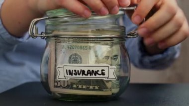 Kadın eli, içi Amerikan doları dolu cam kavanozdan para alıyor. İçinde INSURANCE yazılı nakit para var. Hazırlık para tasarrufu. Orta karar tüketim ve ekonomi. Para toplama