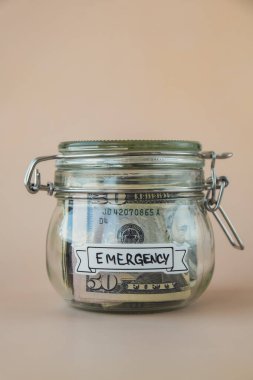 Cam kavanoz dolusu Amerikan doları nakit para ve EMERGENCY yazılı. Hazırlık para tasarrufu. Orta karar tüketim ve ekonomi. Para toplama