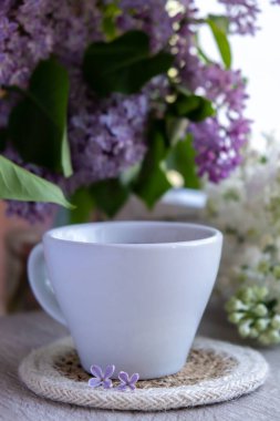 Lezzetli siyah çay beyaz bardakta pencere kenarında aromatik leylak çiçekleriyle. Bahar kompozisyonu bir bardak leylak çayı içerken mor leylak dalları çiçek açıyor. Kopya alanı tebrik kartı için natürmort hayat