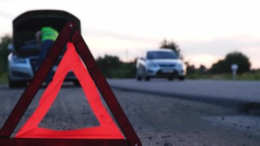 Yansıtıcı yelek giymiş, tanınmaz halde üzgün bir sürücü. Kaputu açık kırık bir arabanın yanında duran erkek sürücü. Kırmızı üçgen diğer yol kullanıcılarını araba arızası ya da motor arızası konusunda uyarmak için kırsal bölgede durur