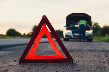 Yansıtıcı yelek giymiş, tanınmaz halde üzgün bir sürücü. Kaputu açık kırık bir arabanın yanında duran erkek sürücü. Kırmızı üçgen diğer yol kullanıcılarını araba arızası ya da motor arızası konusunda uyarmak için kırsal bölgede durur