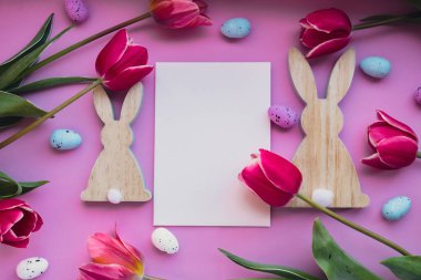 Beyaz boş zarf paskalya yumurtalarını ve menekşe rengi pastel arka planda pembe laleli ahşap tavşanları kopyalıyor. Tebrik kartı. Düz kompozisyon üst görünümü. Dekorasyon minimalist modern tasarım