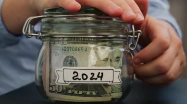 Kadın eli, içinde Amerikan doları olan cam kavanozdan 2024 sayılı nakit banknot alır. Hazırlık para tasarrufu. Orta karar tüketim ve ekonomi. Para toplama