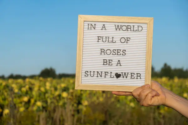 IN A WORLD FULL OF ROSES BE A SUNFLOWER text on white board next to sunflower field. Sunny summer day. Motivational caption inspirational quote. Be unique concept