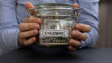 Amerikan dolarlarıyla dolu cam kavanozu elinde tutan tanınmayan bir kadın. İçinde EMERGENCY mesajı olan nakit banknot. Hazırlık para tasarrufu. Orta karar tüketim ve ekonomi. Para toplama