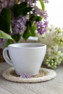 Lezzetli siyah çay beyaz bardakta pencere kenarında aromatik leylak çiçekleriyle. Bahar kompozisyonu bir bardak leylak çayı içerken mor leylak dalları çiçek açıyor. Kopya alanı tebrik kartı için natürmort hayat