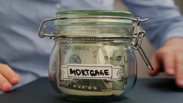 ドル紙幣で満たされたガラスジャーの貯金の前に屋根の印を示す認識できない女性 ジャーの前に写したMortgage 将来のための個人資金の追加収入の管理 — ストック動画