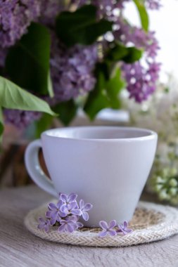 Lezzetli siyah çay beyaz bardakta pencere kenarında aromatik leylak çiçekleriyle. Bahar kompozisyonu bir bardak leylak çayı içerken mor leylak dalları çiçek açıyor. Kopya uzay tebrik kartı, poster, afiş, duvar kağıdı için hala hayat var. Rahatlama ve doğal