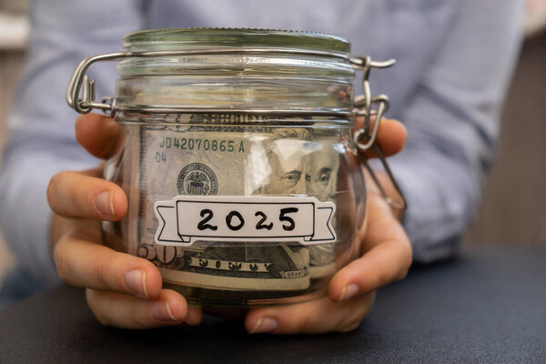 Unrecognizable woman holding Saving Money In Glass Jar filled with Dollars banknotes. 2025 year transcription in front of jar. Managing personal finances extra income for future insecurity background