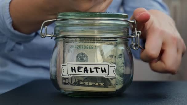 女性的手在装有美元钞票的玻璃瓶中增加了钱 在罐子前的健康转录 为今后的不安全背景管理个人财政额外收入 — 图库视频影像