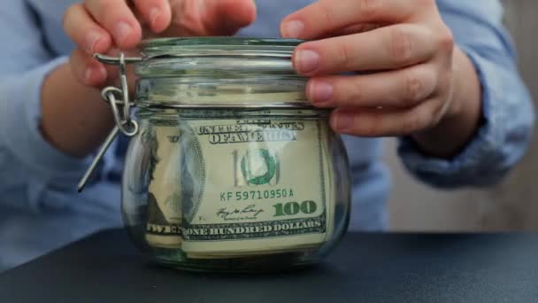 女性手存钱转玻璃瓶装满美元钞票 未来转录在罐子前 为今后的不安全背景管理个人财政额外收入 — 图库视频影像