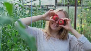 Serada kırmızı domatesleri gözlerine bastıran mutlu sarışın kadın. Yeşil bahçede eğlenen dişi çiftçi mevsimlik organik sebzeleri hasat ederken. Yerel yetişmiş konsept 