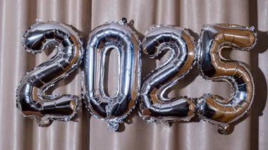 Atlas Tekstil arka planında 2025 Metalik Balon Mutlu Yıllar. Gümüş folyo balonları Noel tatili konseptini temsil ediyor. Kutlama partisi dekorasyonu.
