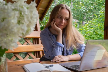 Young happy woman focuses on her laptop in wooden alcove. Relaxed outdoor setting emphasizes comfort and productivity. Remote work learning concept clipart